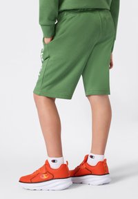 Enfant portant un short vert et des baskets orange avec des chaussettes blanches Champion, se tenant les mains dans les poches contre un fond uni.