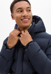 Granatowa kurtka puchowa o pikowanej teksturze, z kapturem i zamkiem błyskawicznym. Kołnierz jest beżowy, co stanowi kontrast dla ciemnego koloru zewnętrznego.