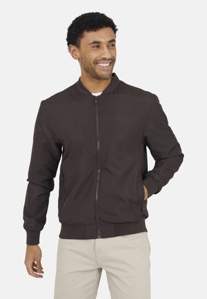 Man die glimlacht, draagt een donkerbruine bomberjack met rits en heeft zijn handen in de zakken van lichtgekleurde broek, staand tegen een effen achtergrond.