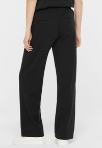 Schwarze weite Hosen mit weicher Textur, ausgestattet mit einem elastischen Taillenbund und zwei Gesäßtaschen. Kombiniert mit weißen Sneakers.