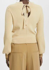 Pull en tricot côtelé beige à manches longues, avec un détail noué au dos du col et une découpe ouverte à l'arrière, associé à un pantalon taille haute couleur camel.
