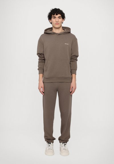 Sweat-shirt marron avec une capuche et une poche frontale, assorti avec un pantalon fuselé, tous deux en tissu doux. Des baskets blanches complètent la tenue.