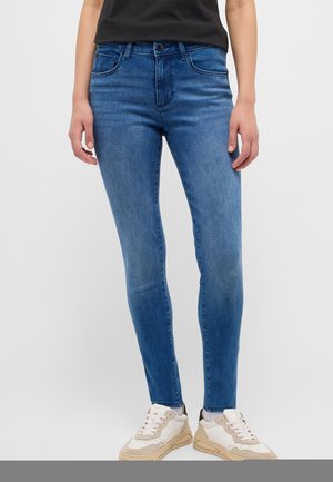 Femme portant un jean slim bleu avec un léger délavage et des baskets blanches, debout devant un fond gris uni.