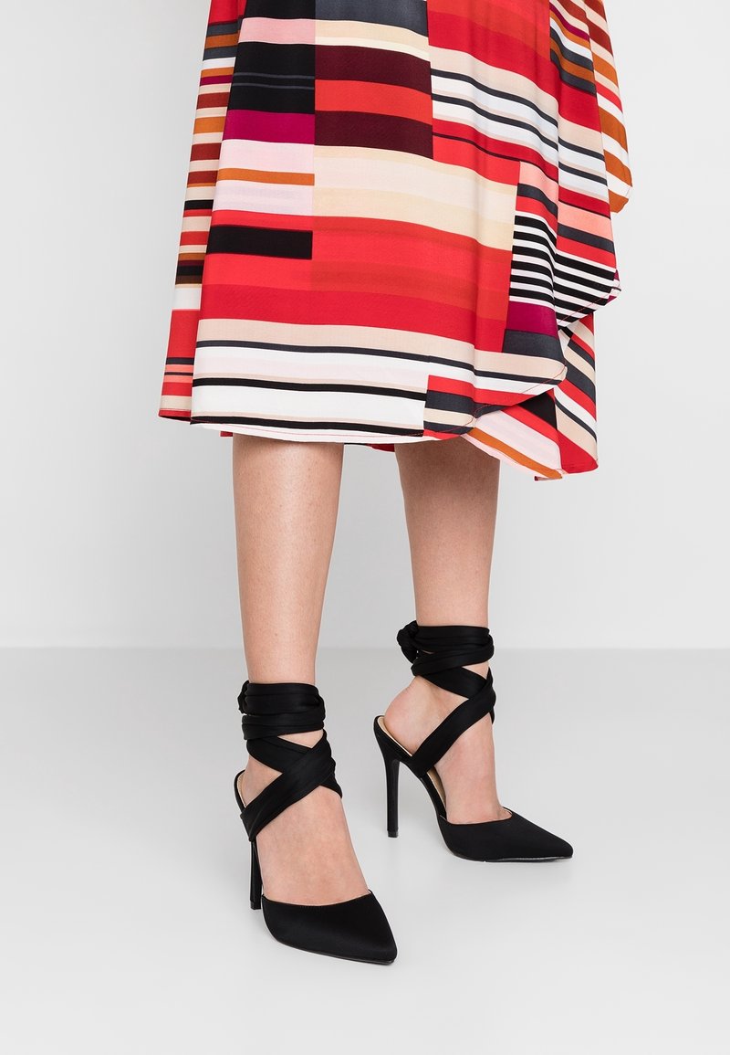 Femme portant des chaussures noires à talons hauts et à brides à la cheville et une jupe mi-longue rayée colorée, debout sur un sol gris clair.