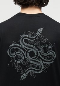 Άτομο με κοντά σκούρα μαλλιά που φοράει μαύρο πουκάμισο με μεγάλο γκρι φίδι σχεδιασμένο στην πλάτη και με στυλιζαρισμένο κείμενο.