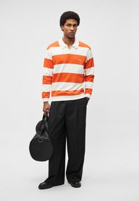 Polo de manga comprida às riscas laranja e brancas com logótipo lateral, combinado com calças pretas largas e uma mala preta redonda com pega.