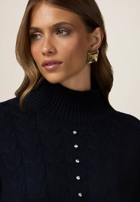 Pulover navy cu tricotaje torsadate, având un guler înalt cu cordon, cu cinci butoni decorativi cu strasuri pe față. Cercei geometrice aurii.