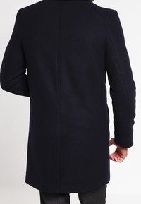 Cappotto in lana blu navy con vestibilità su misura, caratterizzato da una linea dritta, dettagli di cucitura sottili e senza hardware o decorazioni visibili.