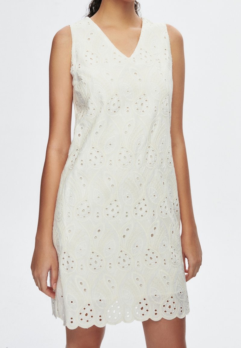 Robe blanche sans manches avec décolleté en V et motif en dentelle avec ourlet festonné, portée par une personne debout sur un fond uni.