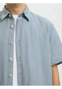 Homme portant une chemise à manches courtes à rayures verticales bleu clair et blanc, boutonnée, par-dessus un t-shirt blanc à col rond.