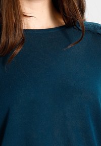 Türkises Strickoberteil mit entspannter Passform, weitem Ausschnitt und dezenten strukturierten Akzenten an den Schultern. Weiche, leichte Stoffqualität.