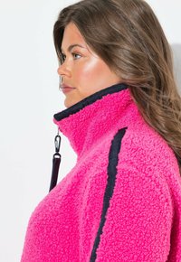 Fleece-takki kirkkaan vaaleanpunainen, jossa on korkea kaulus, mustia yksityiskohtia ja vetoketjukiinnitys. Teksturoitu, pehmeä materiaali, joka näyttää mukavalta.