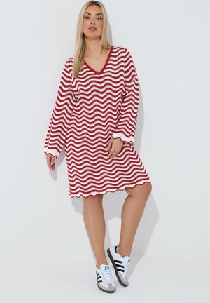 Vrouw staand, dragend een rood-wit golvend gestreepte lange mouw gebreide jurk en witte Adidas sneakers met zwarte strepen.