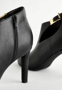 Stivali neri in pelle fino alla caviglia con zip laterale, alti tacchi a stiletto e design a taglio. Accessori in metallo dorato vicino alla zip. Texture opaca.