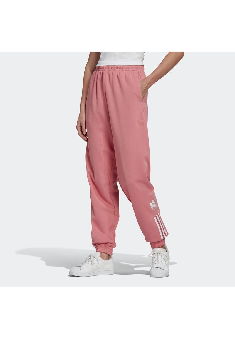 Persoon draagt losse roze Adidas joggingbroek met elastische boorden en witte sneakers, staat met één hand in de zak.