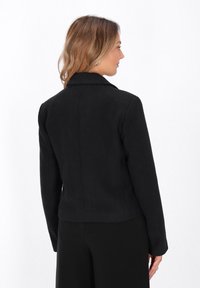 Donna con capelli castani di lunghezza spalla, che indossa un blazer nero aderente e pantaloni neri, rivolta di lato su uno sfondo bianco.