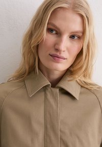 Beżowa kurtka z kołnierzem o wyraźnych detalach szwów, wykonana z fakturowanej tkaniny, z dopasowanym fasonem i minimalistycznym designem.