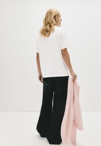 En blond kvinde står med ryggen til iført en løs hvid T-shirt, sorte brede bukser, mens hun holder en foldet lyserød sweater i sin højre hånd.
