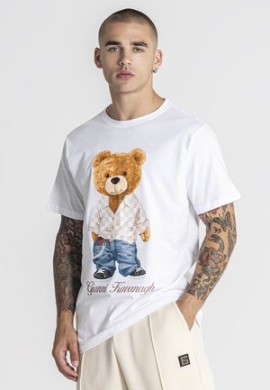 Muž s tetovanými rukami v bielom tričku s obrázkom medvedíka v kockovanej košeli a džínsoch nad textom „Gianni Kavanagh“.