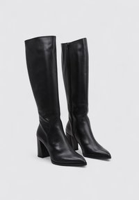 Botas de cuero negro hasta la rodilla con punta afilada y tacón ancho. Cuentan con una cremallera lateral para facilitar su uso. Textura smooth, diseño elegante.