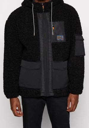 Schwarze Fleecejacke mit Kapuze, Frontreißverschluss, Kordeln, zwei großen unteren Taschen mit Klappen, Brustreißverschlusstasche und einem Aufnäher am linken Arm.