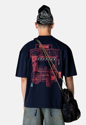 Person iført en mørkeblå skjorte med et rødt grafik af en nudelvogn, lyseblå jeans, en sort hue med hvidt design, og bærer en sort skuldertaske.