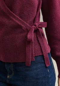 Maglione avvolgente in un profondo tessuto burgundy, caratterizzato da una superficie testurizzata e dettagli con cintura annodata in vita, abbinato a jeans scuri.