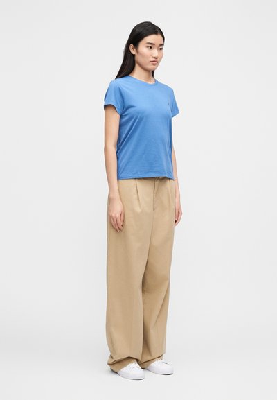Kék pamut rövid ujjú póló, bézs színű, bő szárú nadrággal párosítva. A modell egy egyszínű háttér előtt áll, fehér sportcipőt viselva.