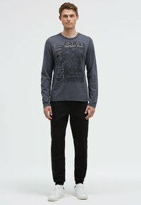 Langärmliges graues T-Shirt mit Grafikdruck, kombiniert mit schwarzen schmalen Hosen und weißen Turnschuhen, vor einem neutralen Hintergrund stehend.