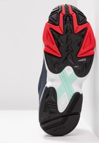 Svart och röd sula på en sneaker med en texturerad design, Torsion-accent i mintgrön, och ett övervägande mörkblått ovandel.