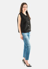Chaleco sin mangas negro con tela texturizada, botones frontales y bolsillos laterales, combinado con jeans ajustados de color azul claro y tacones negros.