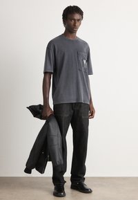 Mann iført en mørkegrå oversized T-skjorte, svarte jeans og svarte sko, holder en svart jakke i venstre hånd, og står mot en ensfarget bakgrunn.