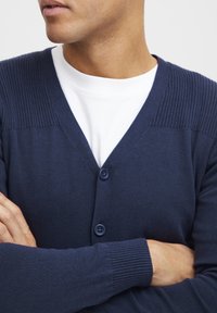 Marineblå cardigan med ribbet skulderdesign, tre knapper og myk strikketekstur, båret over en hvit t-skjorte.