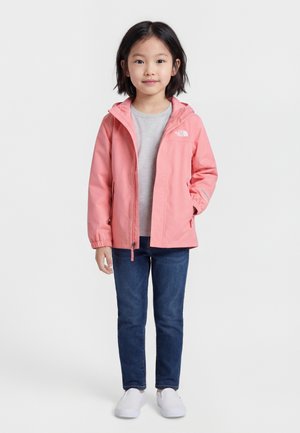 Dieťa v ružovej bunde s kapucňou, sivej košeli, modrých džínsoch a bielych slip-on topánkach, stojace pred jednoduchým pozadím.