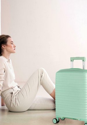 Maleta rígida de color verde menta con superficie texturizada, asa retráctil y cuatro ruedas, situada junto a una persona sentada.