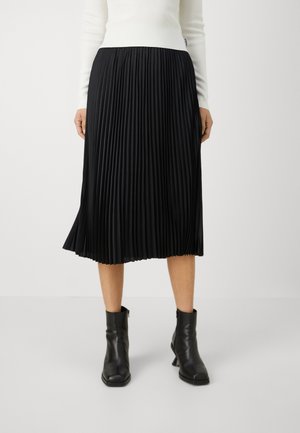 Woman wearing a black pleated midi skirt, white long-sleeve top, and black ankle boots, standing against a plain background.