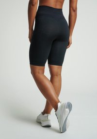 Sportovně založená osoba v černých vysokých cyklistických šortkách a bílých teniskách, stojící s přehnutýma nohama na jednobarevném pozadí.