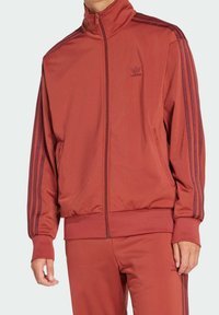 Mörkröd zip-up träningsjacka med hög krage och mörkröda accenter. Har framfickor och tre ränder ner längs ärmarna.