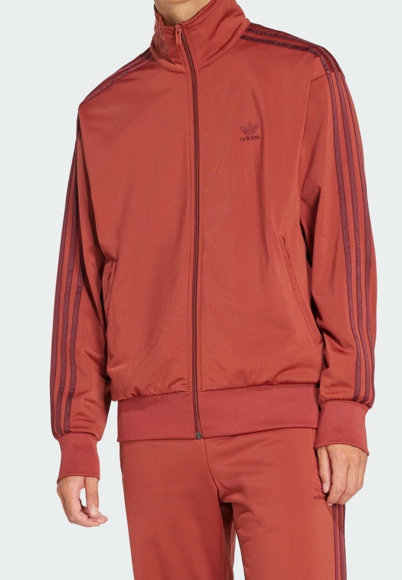 Mörkröd zip-up träningsjacka med hög krage och mörkröda accenter. Har framfickor och tre ränder ner längs ärmarna.