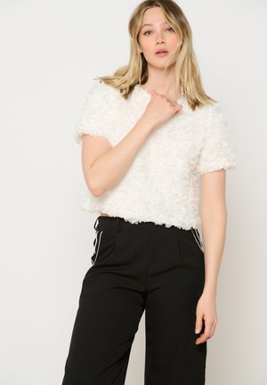White faux fur crop top with short sleeves, featuring a textured floral pattern. Paired with black trousers with white side stripes.