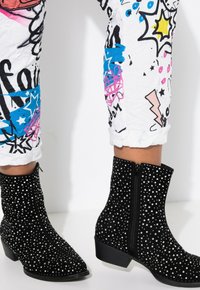Schwarze Wildleder-Ankle-Boots, bedeckt mit silbernen Nieten, mit einem seitlichen Reißverschluss und einem klobigen Absatz, kombiniert mit bunten, gemusterten, cropped Hosen.