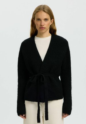 Jonge vrouw draagt een zwarte, pluizige, met ceintuur versierde cardigan over een wit overhemd en lichte broek, staand tegen een effen achtergrond.