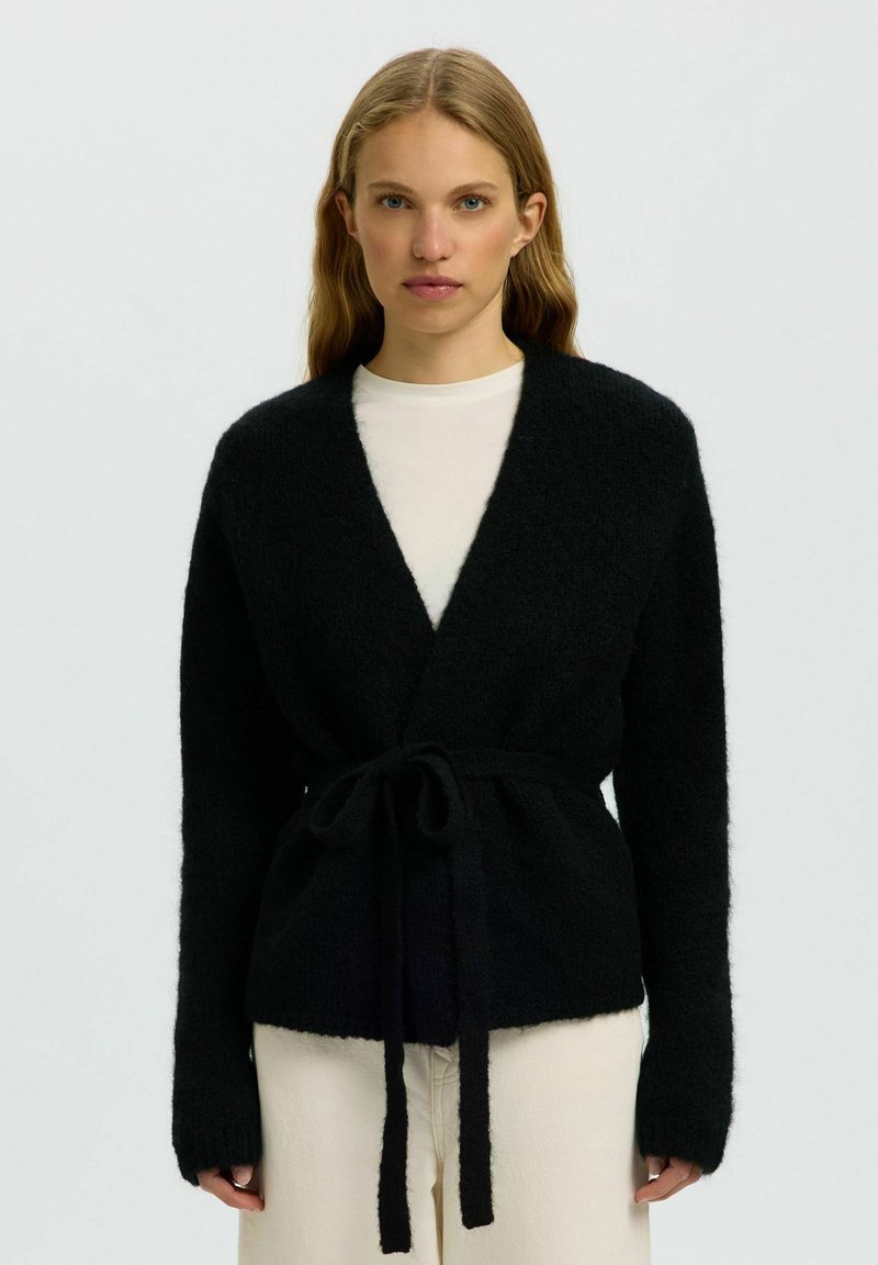 Jeune femme portant un cardigan noir duveteux ceinturé sur une chemise blanche et un pantalon clair, debout devant un fond uni.