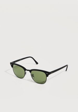 Gafas de sol con montura negra y lentes teñidas de verde, con almohadillas metálicas en la nariz y patillas curvadas sobre un fondo blanco.