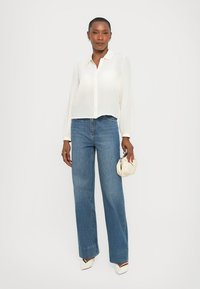 Frau in weißer Bluse mit langen Ärmeln, weit geschnittenen blauen Jeans, weißen High Heels, die eine kleine weiße Tasche hält und vor einem schlichten Hintergrund steht.