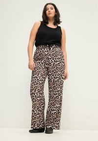 Leopardenmuster, hoch taillierte, weit geschnittene Hosen mit glatter Textur. Ein schwarzes, ärmelloses Oberteil rundet den Look ab. Die Schuhe sind schwarz.