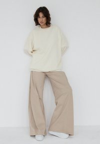 Przewymiarowany kremowy sweter z opuszczonymi ramionami, zestawiony z szerokimi beżowymi spodniami o luźnym kroju. Model nosi białe trampki. Prosty, minimalistyczny styl.