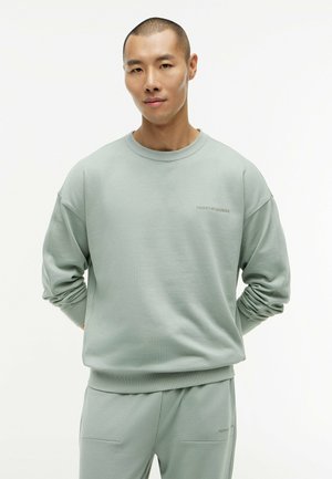 Mann mit kurzen Haaren, der einen hellgrauen Tommy Hilfiger Pullover und passende Hose trägt, steht mit den Händen hinter dem Rücken.