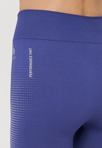 Tesne prilegajoče vijolične legice iz funkcionalnega pletenega materiala. Imajo teksturiran vzorec na eni strani in logo natisnjen blizu pasu.