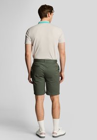 Mand står med ryggen til, iført en beige polo med turkis krave, olivengrønne shorts, hvide sokker og hvide sneakers.